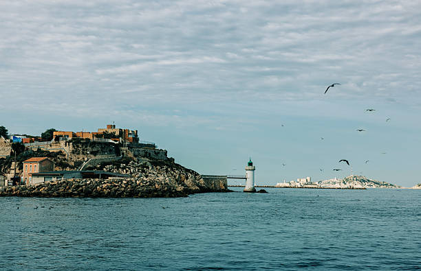 Rocky coastal fortress with lighthouse and flying seagulls facing the sea under cloudy sky near Marseille