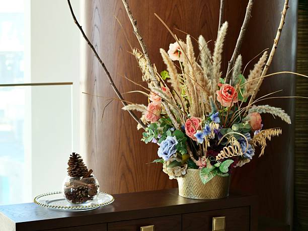 A vibrant, mixed artificial flower arrangement in a textured gold-colored pot, featuringpink roses, blue hydrangeas, purple flowers, and dried grasses (pampas* *grass) on adark wood surface, for interior decor