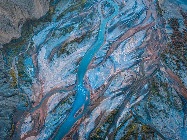 Aerial view of a turquoise river flowing through a rugged desert landscape in Xinjiang, China. Braided channels carve through sediment layers and arid terrain, creating striking natural patterns and color contrasts seen from above.