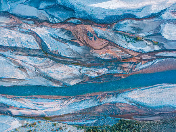 Abstract aerial view of a river flowing through a textured landscape in Xinjiang, China. Layers of blue water, pale sediment, and earth-toned deposits form painterly patterns and natural lines, creating a visually striking composition seen from above.
