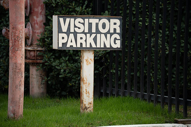 A weathered visitor parking sign stands near a rusty industrial pipe and a black metal fence, surrounded by lush green grass and foliage
