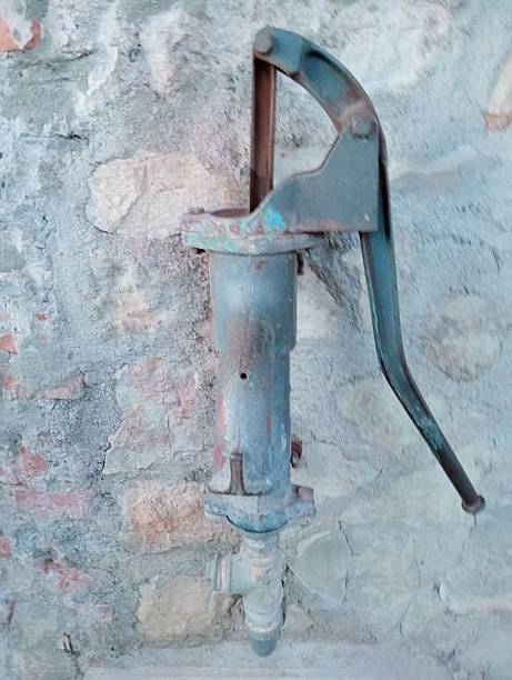 A close-up of an antique, rusty manual water pump installed on a traditional rustic stone wall. This weathered mechanical tool represents historical irrigation and traditional rural life. Ideal for themes of history, agriculture, and vintage technology