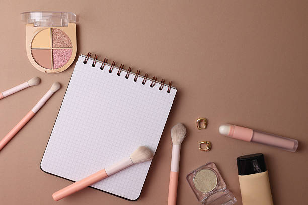 Top View Cosmetics Layout With Brushes, Palettes, Sponge, Lipstick And Notebook Arranged On Soft Beige Surface