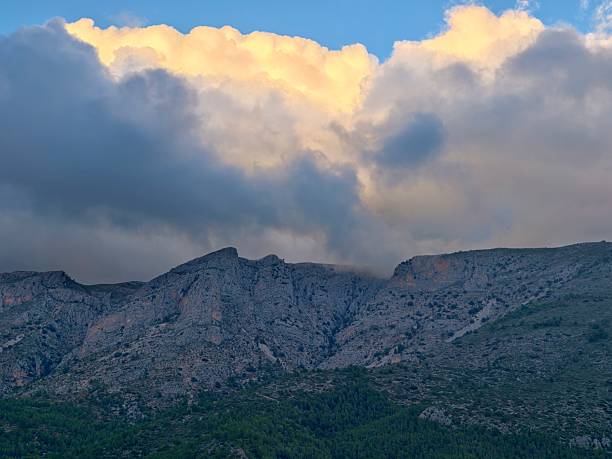 Sunlit Mountain Range Under Glowing Cloud Crown, Soft Golden Highlights Brush Rocky Ridges And Distant