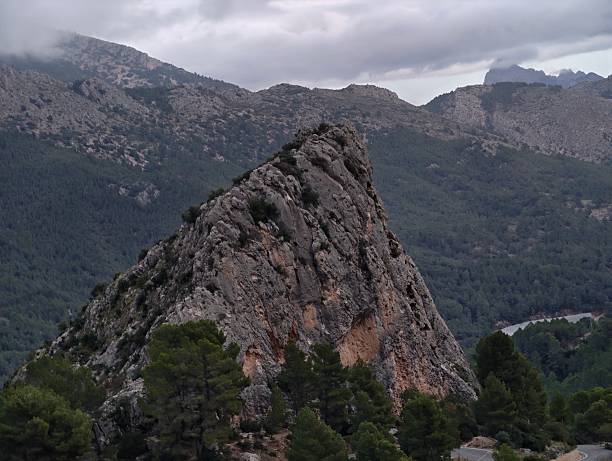 Jagged Rock Spire Rising From Valley, Exposed Strata And Textured Cliff Face Framed By Pine Foothills