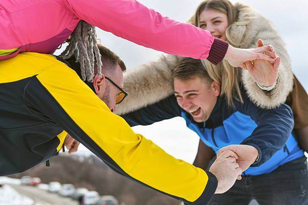 A group of friends enjoy a playful moment in a snowy landscape, linking hands and leaning toward each other with laughter. Colorful jackets, winter scene, energy, and camaraderie.