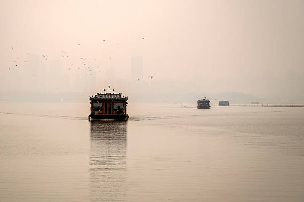 A single passenger boat creates a perfect reflection on calm East Lake, with birds flying through the hazy sky in Wuhan
