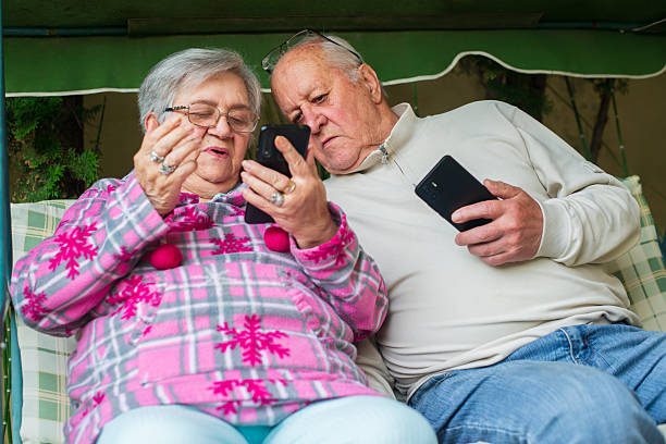 Senior couple sitting in the back yard in the porch swing in summer, using smart phones.