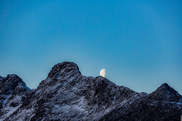 Svolvear, Lofoten Island, Norway Large moon is rising over a mountain range. The sky is clear and blue, and the mountains are covered in snow. The scene is peaceful and serene, with the moon casting a soft glow over the landscape
