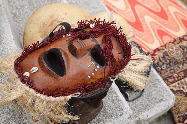 Wooden mask from africa with raffia beard and hair, cowrie shells, and red decorative string, resting on a small drum