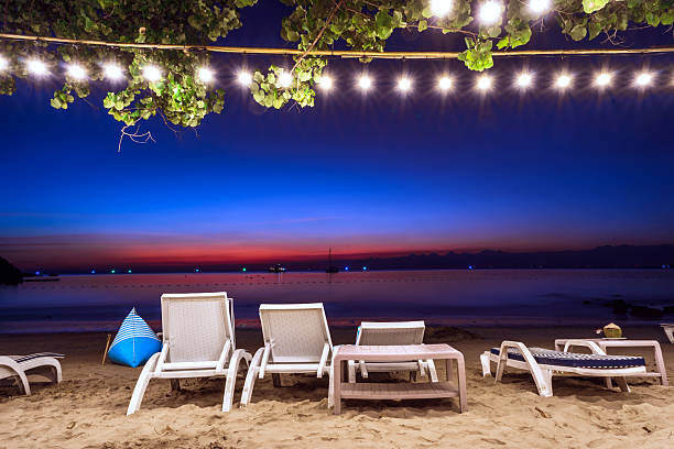 Relaxing beachfront lounge at night with white chairs, decorative string lights, and a vibrant blue and orange twilight sky, Koh Samed, Thailand