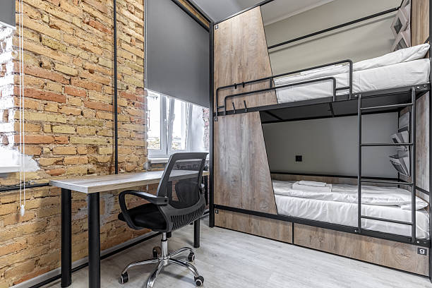Interior view of a hostel dormitory featuring a black metal bunk bed with wood privacy panels and integrated storage. The room includes a wooden desk with a black mesh office chair and a rustic exposed brick accent wall.
