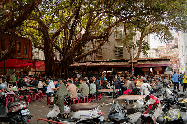 Taipei, Taiwan - March 13, 2024: Vibrant outdoor lunch scene in the temple courtyard outside Dadaocheng Cisheng Temple, where locals gather around long tables under a massive ancient banyan tree. The wide-angle view captures the lively atmosphere with numerous diners enjoying meals amid parked scooters and traditional architecture. Red lanterns and temple structures frame the bustling communal dining area.