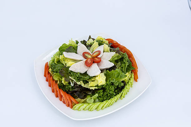 Studio shot of chicken breast salad with mixed leafy greens, sliced cucumber and tomatoes, served on a white plate against a white background.