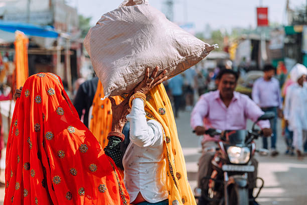 Colorful street market in Pushkar, Rajasthan shows locals in traditional attire. A woman carries a large bag on her head while a man rides a bike through a busy market alley.