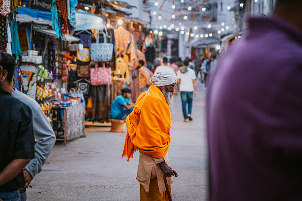 A saffron-clad Man walks through a busy street market in Pushkar, Rajasthan. Colorful stalls, string lights, and shoppers create a vibrant scene blending culture, spirituality, and daily life.