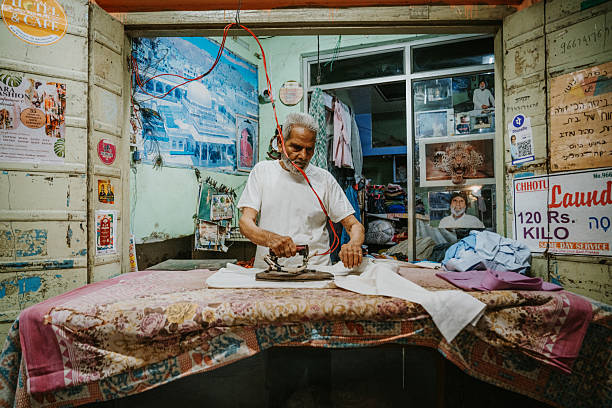 A seasoned artisan irons fabric in a small Pushkar shop, surrounded by posters and fabrics. A glimpse into traditional Indian laundry work and local commerce, full of color, texture, and daily craft.