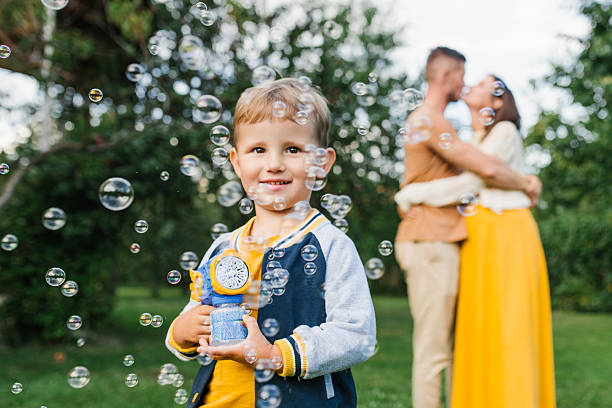 Young boy enjoying playtime with bubble machine outdoors, smiling happily while parents embrace in background. Image symbolizes family love, happiness and joyful childhood.