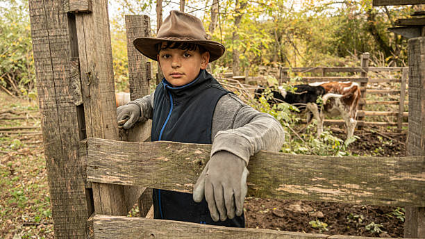 Concerned young cowherd wearing a brown hat and leaning on a wooden fence, observing the rural farm landscape and other people working in the background