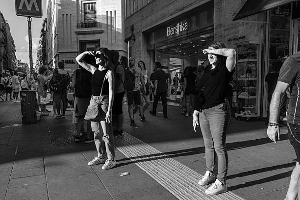 NAPLES, ITALY - June, 2024, 2022: Pedestrians stand on the sidewalk of Via Toledo, the city's main shopping street, shielding their eyes from the harsh afternoon sun. In the background, modern storefronts like \