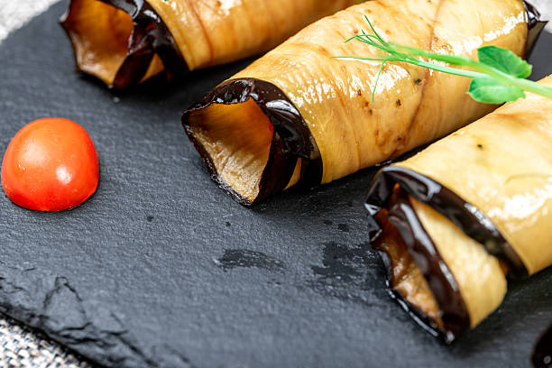 Delicious traditional Georgian appetizer, Badrijani, consisting of fried eggplant slices rolled with a flavorful filling (likely walnut or cheese/garlic paste) and garnished with fresh microgreens and cherry tomatoes. Served on a modern black slate board against a textile background. Focus on healthy, organic, and vegan food.