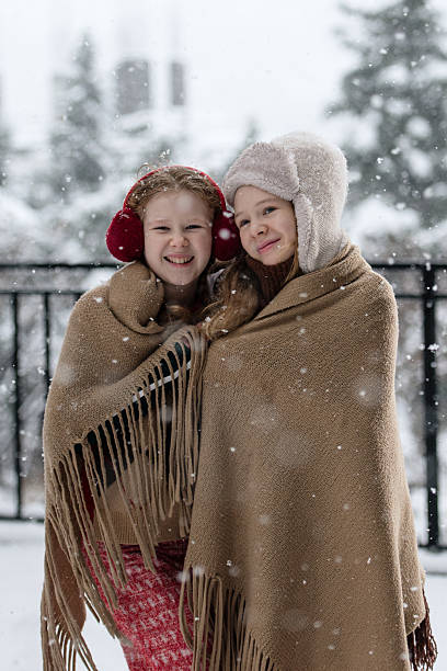Portrait of two sisters hugging under a shared blanket while snow falls around them on a cold winter day.