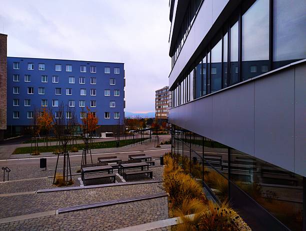 A contemporary blue residential or academic building situated near Altes Landgut in Vienna. The shot features sleek glass facades with reflections and an organized outdoor seating area with benches.