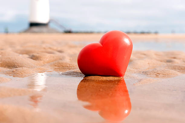 A red heart shape rests on wet sand at the beach. In the background, a lighthouse stands. The tide is low, creating a reflective surface around the heart.