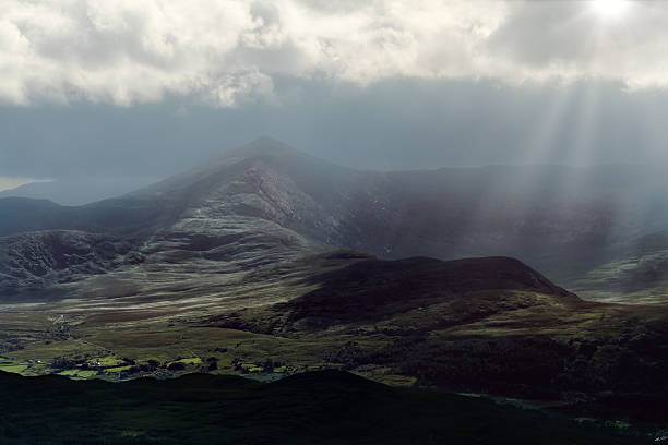 Sunbeams pierce dramatic clouds over rugged mountain slopes, casting light across a tranquil valley.