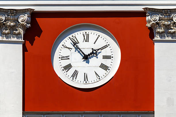 Round wall clock with Roman numerals mounted on building facade. Decorative capitals and architectural details visible around clock.