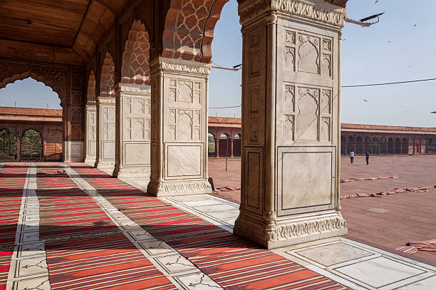 Delhi, India - March 29, 2024: The arcaded walkway of Jama Masjid mosque is shown with white marble columns featuring carved panels, red sandstone arches, striped prayer rugs on the floor, and a view of the open courtyard with a few people in the distance under a clear sky.