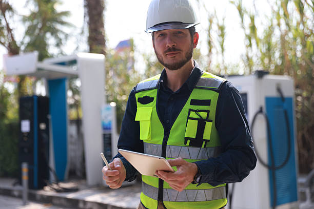 Portrait EX 26-04-01-5 An. industrial engineer or electrical engineer  wearing a hard hat and high-visibility vest uses a tablet at an electric vehicle charging station outdoors