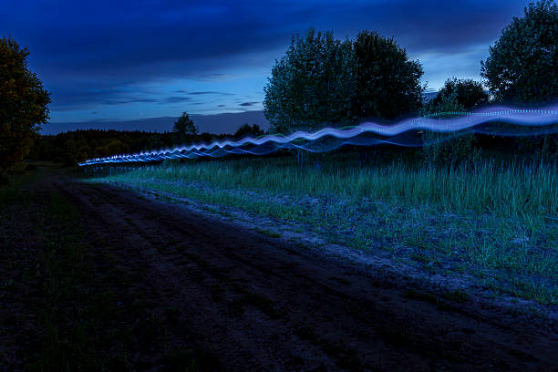 Long exposure shot of a glowing light trail over a dirt road at night in a rural landscape. Dark trees and fields create a quiet, mysterious atmosphere