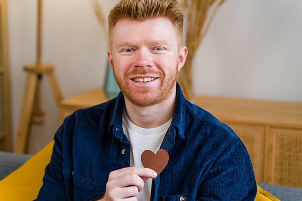 Happy ginger man holding a heart shape on chest, expressing positive emotions indoors