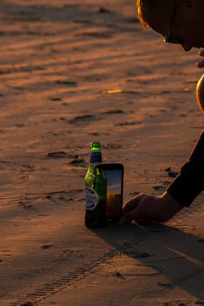 Tofino, Vancouver Island, 27 July 2025A person stands on the sand at sunset in Tofino, Vancouver Island, wearing black sneakers and shorts. A green Peroni beer bottle and a smartphone are placed upright nearby, with the phone screen displaying a live view of the beach and golden sky. The warm light and long shadows evoke a peaceful moment of relaxation and connection with nature.