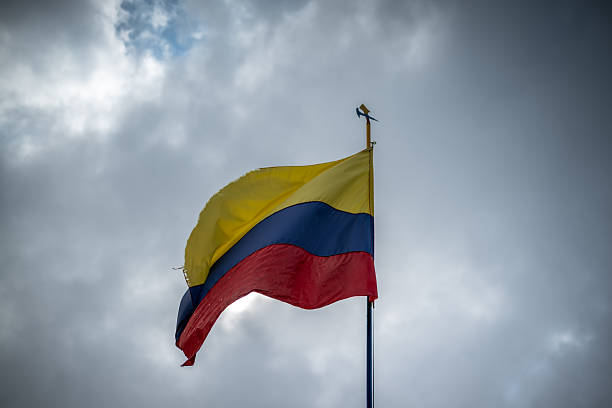 A worn and damaged Colombian flag flies on a flagpole under a cloudy sky in Colombia. The image visually documents the national flag in deteriorated condition, which may symbolize uncertainty, crisis, or challenging moments in the country’s social, political, or economic context.