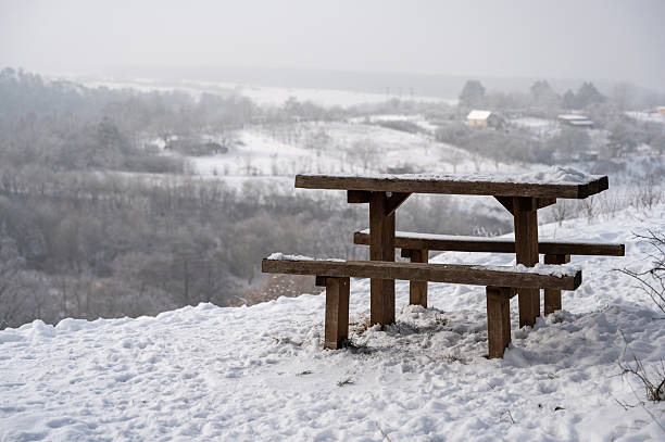 A snow-covered wooden picnic table stands at a quiet winter viewpoint overlooking a misty valley. The soft, frosty landscape and muted colors create a peaceful and atmospheric winter scene.