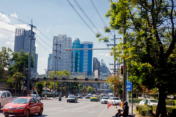 Traffic on Phetchaburi Road at junction to Sukhumvit Rd 3 at lunchtime in Bangkok. View to west. A footbridge of over street. In background are apartment towers and hotels. At bottom right people are on motorcycle