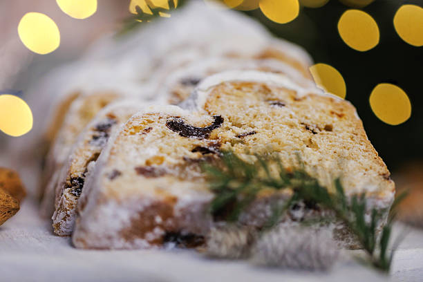 A close-up of sliced Christmas stollen bread dusted with powdered sugar, surrounded by almonds, pinecones, and blurred golden festive lights, evoking a warm holiday atmosphere.