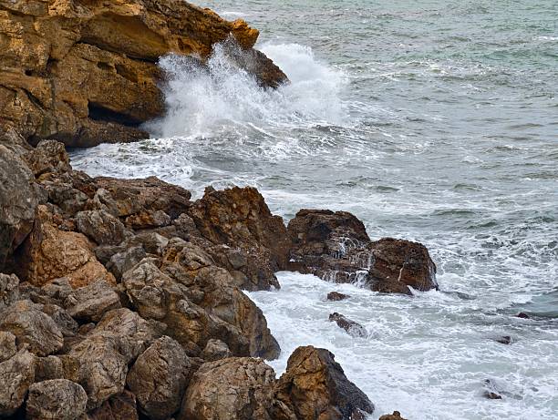 Rocky Coast Waves Crashing Into Shore, CloseUp Surf Hitting Jagged Boulders, White Foam Spray And Turbulent