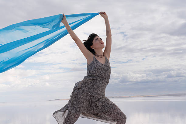 A Caucasian young female in a white sleeveless top and blue jeans stands on a sand dune in the Dubai desert with out-of-focus dry grass in the foreground and clear blue skies above.