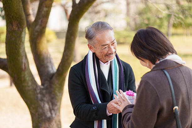 A series of images showing an elderly man presenting a small gift box to a woman while standing outdoors in a public park during daytime. The woman is receiving the gift and looking at it with appreciation as they face each other. The scene shows two senior adults interacting in a natural park environment with trees and open space in the background, representing gift giving, surprise, companionship, and emotional connection, with copy space visible.