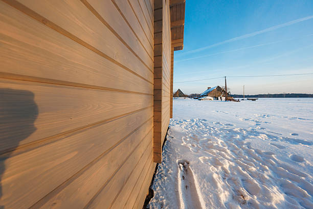 Side view of modern wooden facade faces a snow field with footprints, utility poles, and rustic outbuildings as low afternoon light casts long shadows in a rural setting.