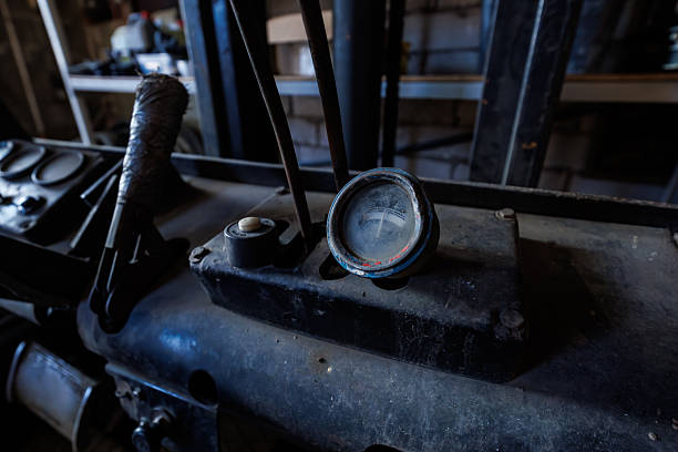 Close view of a weathered round gauge with a chipped blue rim on a grime coated forklift dashboard, beside rusted levers and a taped gear stick in an indoor workshop.