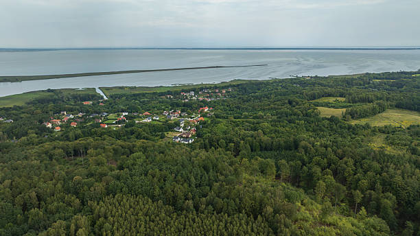 Aerial view shows red and white roofs in dense forest by a calm lagoon. A long breakwater spans the water as a channel cuts inland under soft overcast light.