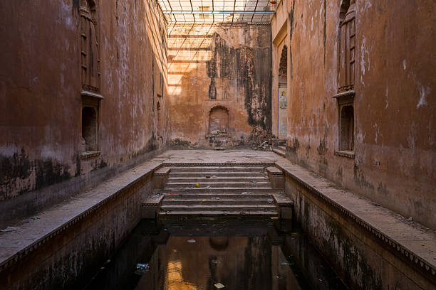 Bundi, India - March 19, 2022: A rectangular stone stepwell with high weathered walls, arched niches, and a metal grid roof overhead, with dark water at the bottom and debris scattered around the steps.