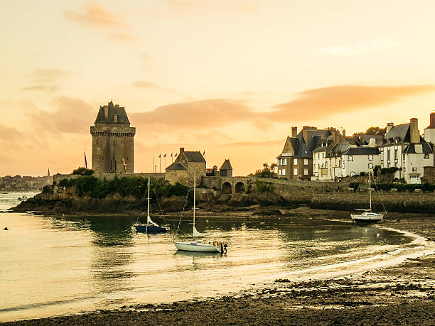 Sailboats float in calm harbor waters at sunset beneath historic ramparts and waterfront houses in Saint-Malo, Brittany, capturing the golden light of a summer evening on the French coast.