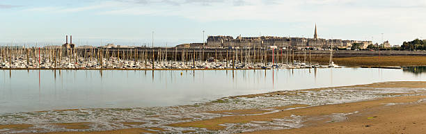 Wide view of the marina in Saint-Malo, Brittany, with numerous sailboats and yachts docked. In the background, the iconic granite buildings and slate roofs of the walled historic city (Intra-Muros) skyline, under a bright sky.
