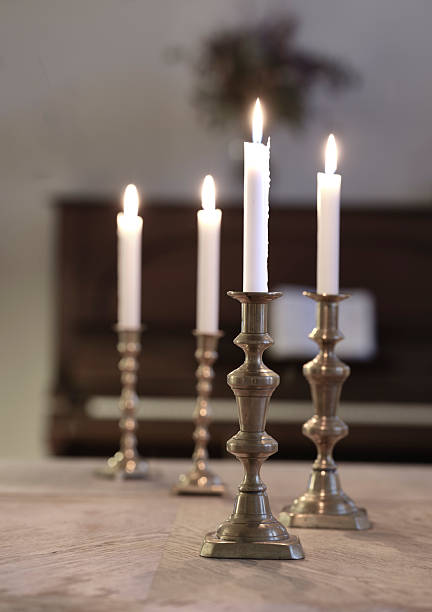 A serene still life features three or four silver candlesticks holding white candles, casting a gentle, warm glow over a rustic wooden table. The scene exudes calm, coziness, and a classic dining ambiance, with subtle reflections and a timeless, Nordic aesthetic that hints at Danish design influences.
