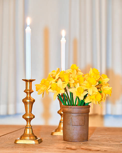 A warm, candlelit still life featuring four brass candlesticks with tall white candles arranged on a wooden dining table. Soft bokeh hints at a piano and indoor greenery in the background, evoking comfort, quiet evenings, family gatherings, and festive ambience.